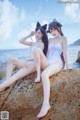 Two women in white bathing suits sitting on a rock by the ocean.